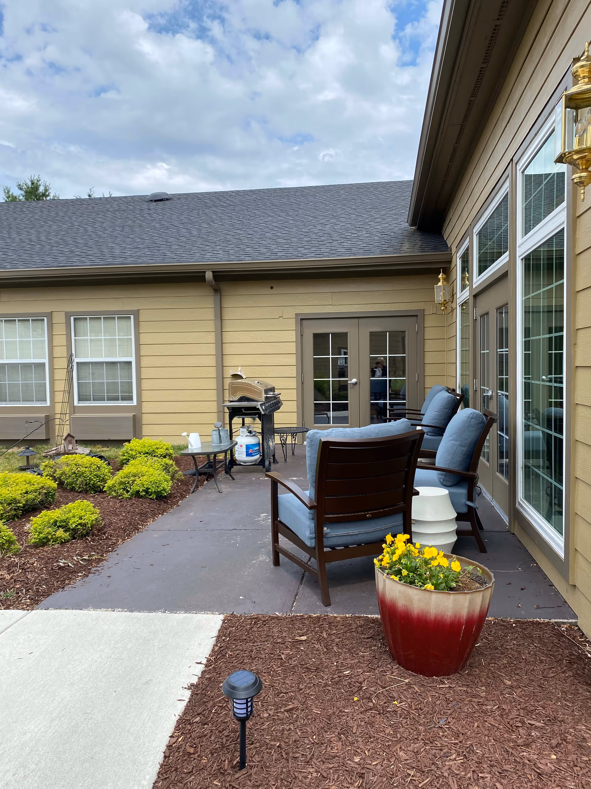 Outdoor patio area at American House Brentwood with cushioned wooden chairs, a small table, a barbecue grill, and a large flower pot with yellow flowers. The patio is adjacent to a beige building with multiple windows and glass doors. There are small bushes and mulch landscaping around the patio.