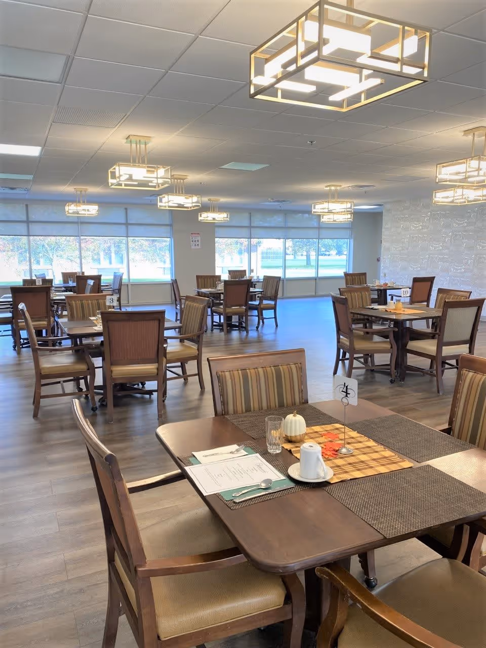 A spacious dining room with multiple wooden tables and chairs arranged neatly. Each table has placemats, menus, and some have small decorative items like a white pumpkin and autumn leaves. Large windows along one wall let in natural light, and modern square pendant lights hang from the ceiling.