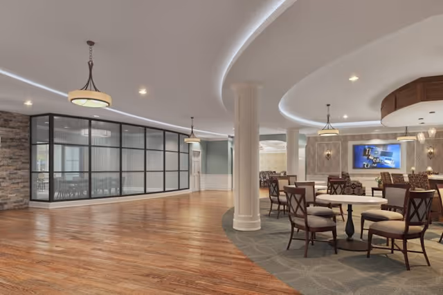 Spacious senior living facility common area with wooden flooring and carpeted seating area. Round tables with cushioned chairs are arranged near a wall-mounted TV. The ceiling features modern lighting fixtures and recessed lighting. A glass-walled room is visible on the left side, and decorative columns are present throughout the space.