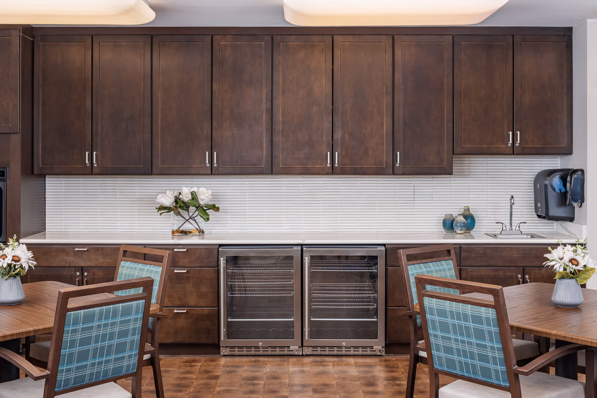 A kitchen area with dark wooden cabinets, a white tiled backsplash, two glass-door refrigerators, a small sink with a faucet, and a wall-mounted paper towel dispenser. In front of the kitchen counter are wooden tables with chairs that have blue plaid cushions. There are flower arrangements on the tables and countertop.