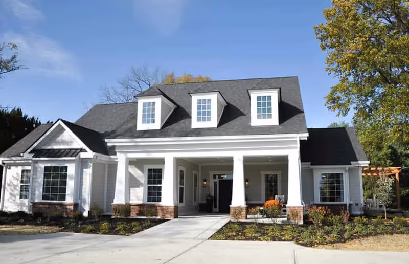 White cottage-style senior living facility front entrance with columns, dormer windows, and landscaped walkway.