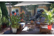 Outdoor patio area with wicker furniture including chairs and a loveseat, a round coffee table, various potted plants, and a large umbrella providing shade. The setting is surrounded by greenery and flowers with a building visible in the background.