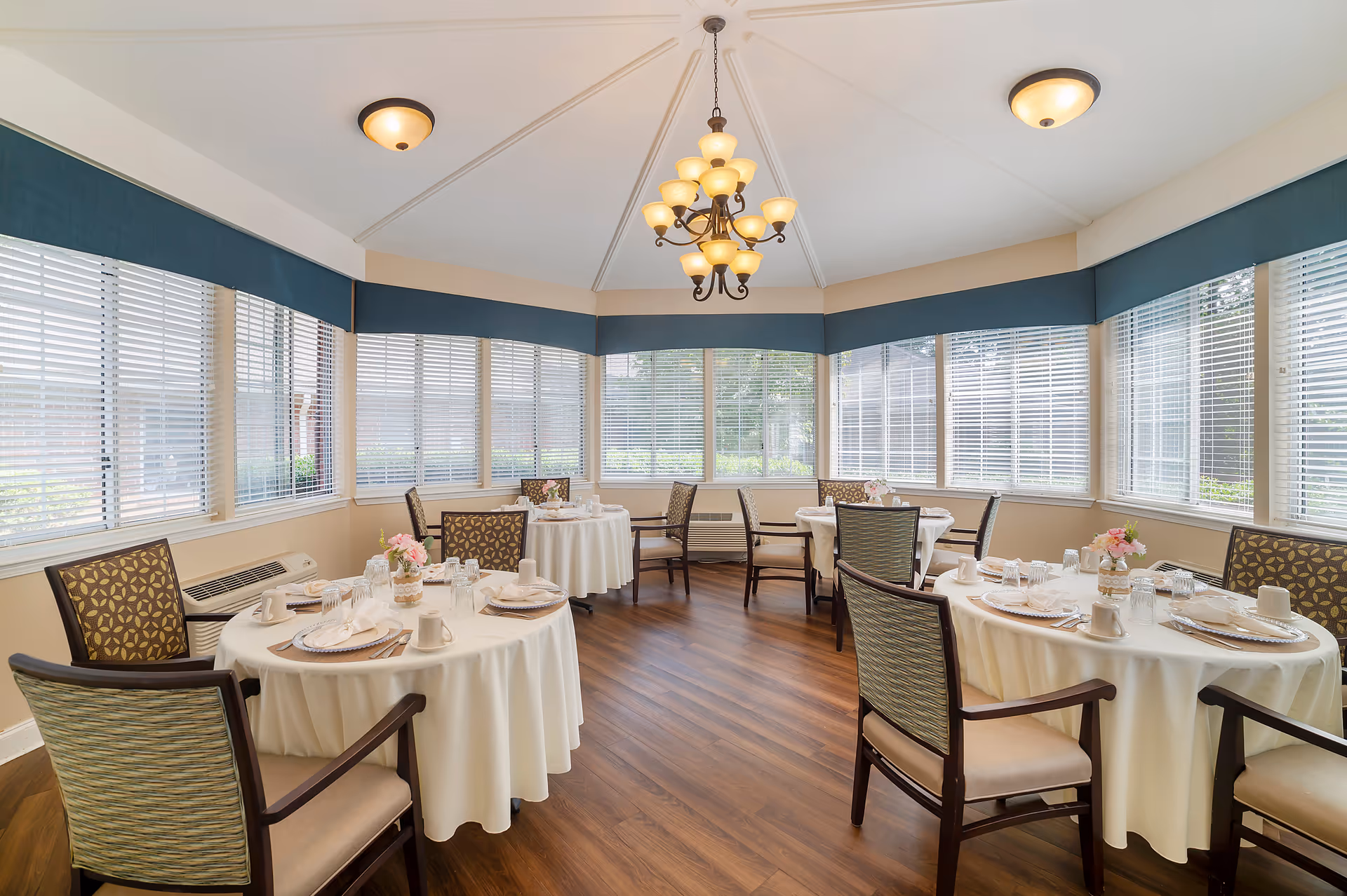 Bright dining room with round tables set for meals, upholstered chairs, large windows with blinds and a central chandelier.