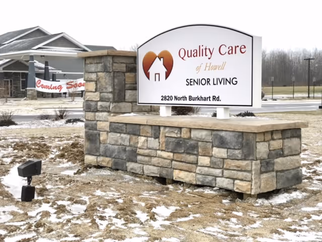 Stone entrance sign reading 'Quality Care of Howell SENIOR LIVING' in front of the facility with the building and a 'Coming Soon' banner visible in the background.