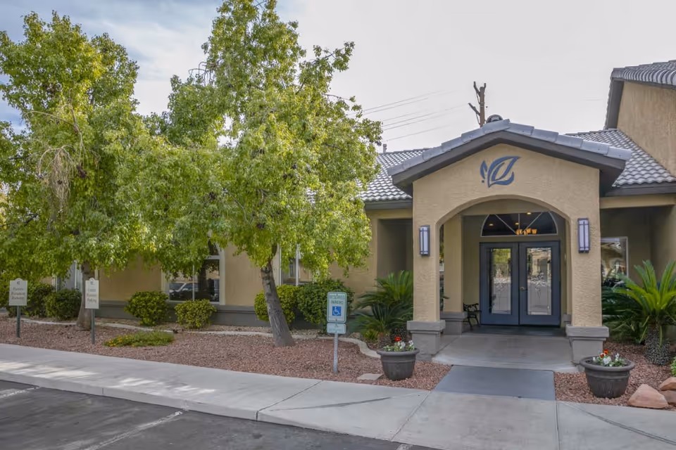 Front entrance of Villa Court Assisted Living showing an arched entryway with double glass doors, potted plants, and trees.