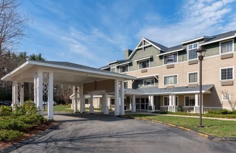 Exterior view of Ledgewood Bay Assisted Living facility showing a multi-story building with beige and green siding, multiple windows, a covered driveway entrance, and a lamppost on a sunny day with a partly cloudy sky.