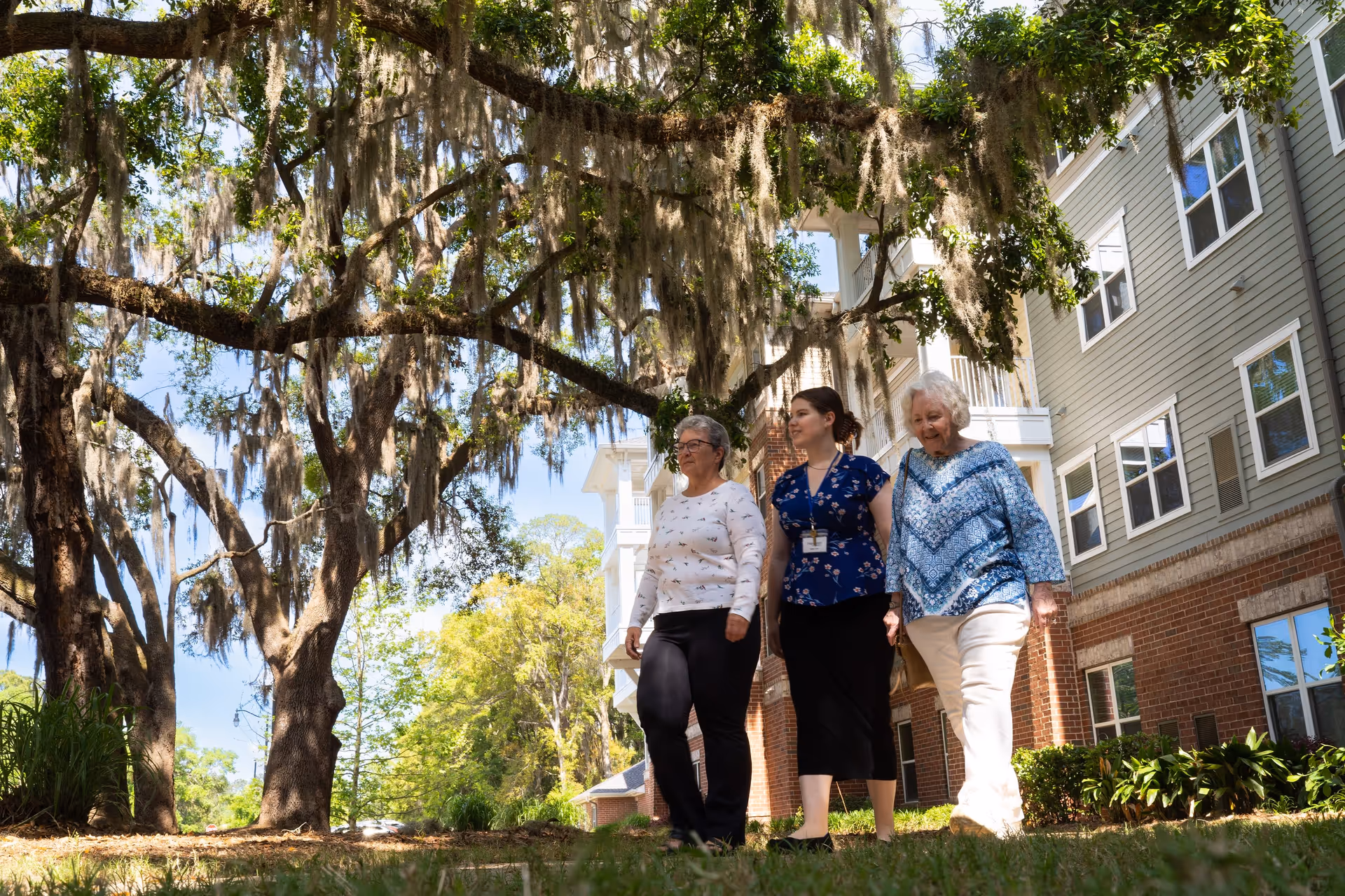 Three women walking outside near a senior living facility building with large trees draped in Spanish moss overhead. The building has gray siding with white trim and brick accents. The women appear to be enjoying a sunny day.