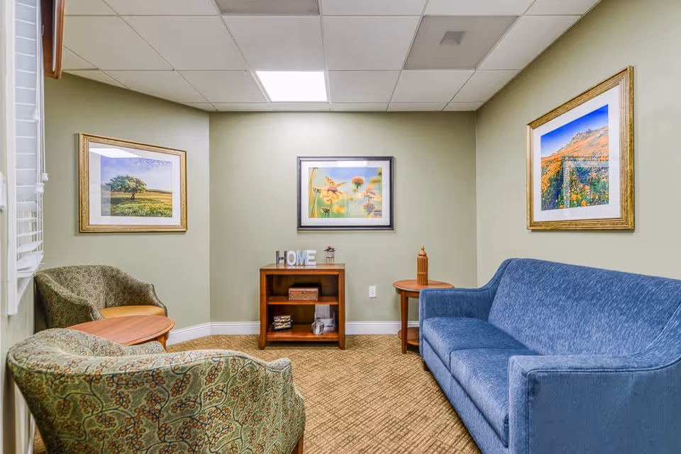 A cozy sitting area in a senior living facility with a blue sofa, two patterned armchairs, a small wooden bookshelf with decorative items including a 'HOME' sign, and three framed landscape photographs on light green walls.