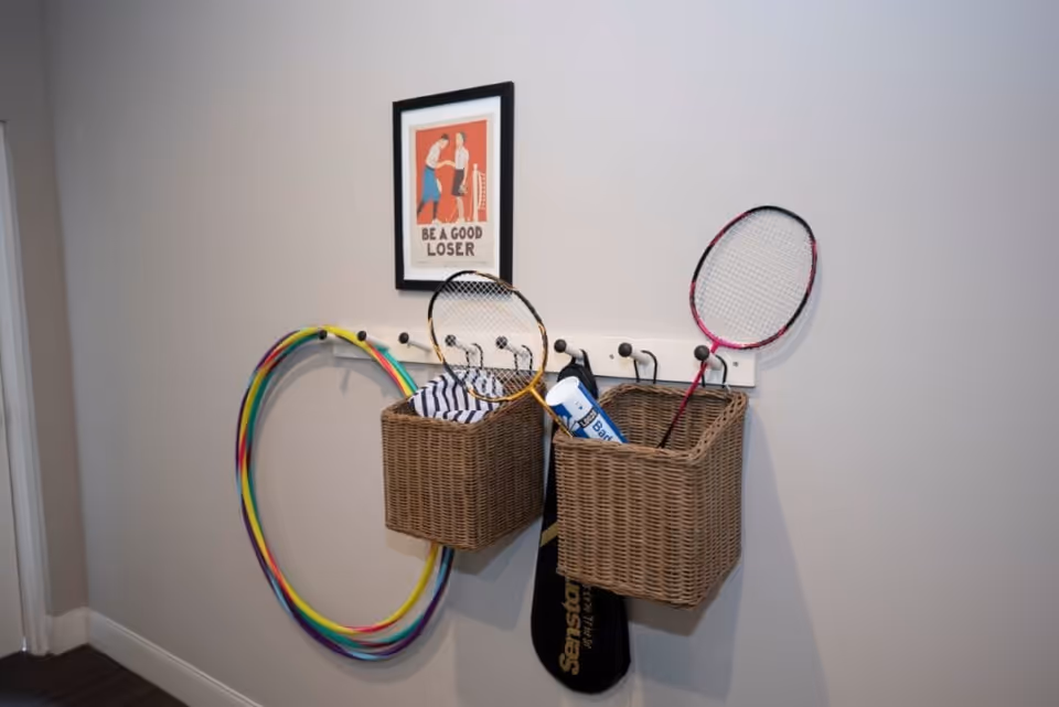Two wicker baskets hanging on a wall-mounted rack with hooks, containing badminton rackets and shuttlecocks. Two colorful hula hoops are hanging on the left side of the rack. Above the rack is a framed poster with an illustration of two people playing badminton and the text 'BE A GOOD LOSER'.