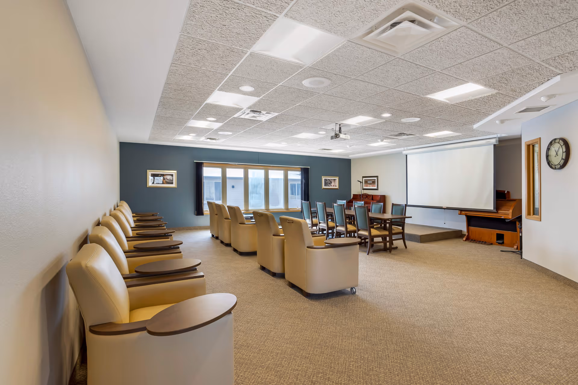 A spacious meeting or activity room with rows of beige armchairs with attached wooden trays on the left side and a set of wooden chairs around tables in the center. The room features a large window with dark curtains, a projector mounted on the ceiling, a projection screen, a piano, and a wall clock. The walls are painted in neutral tones with some framed pictures.