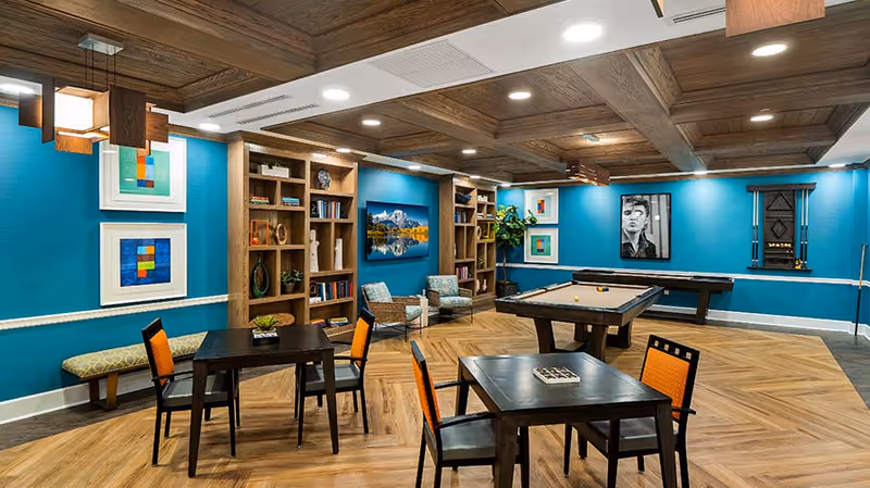 A recreational room with teal walls and wooden coffered ceiling featuring two pool tables, several black tables with orange and black chairs, built-in wooden bookshelves, framed artwork on the walls, and a bench along one wall.
