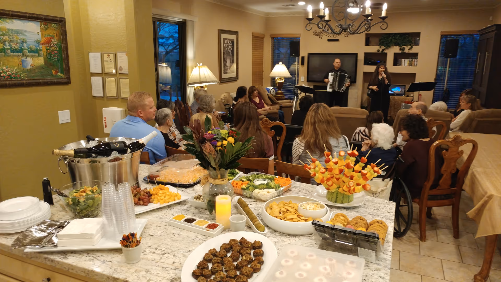A group of people seated in a living room area watching a musical performance with a man playing an accordion and a woman singing. In the foreground, there is a table with various snacks, including fruit skewers, crackers, cookies, salad, and drinks. The room is warmly lit with lamps and a chandelier.