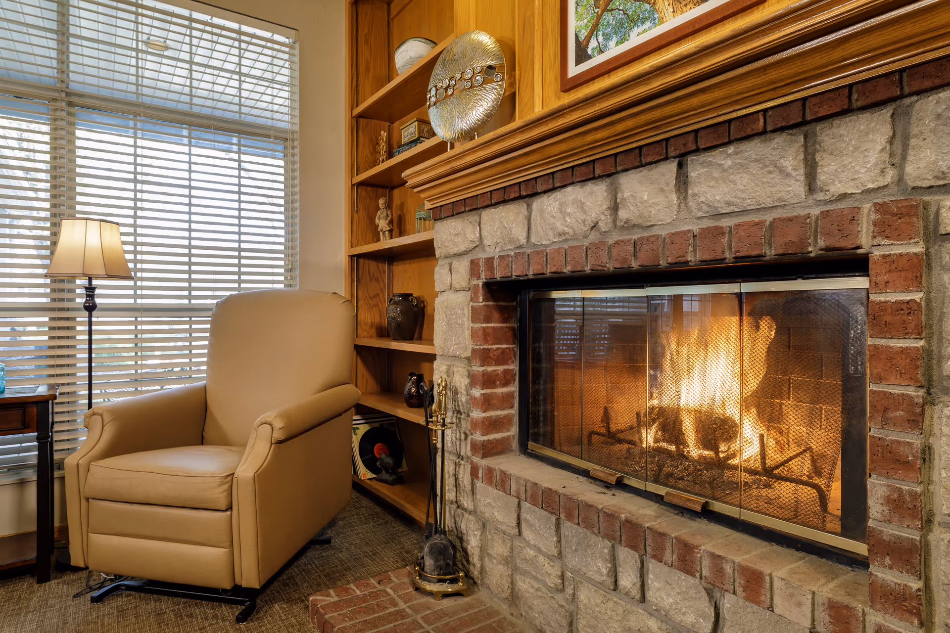 Cozy living area with a beige armchair next to a window with blinds, a floor lamp, a wooden bookshelf with decorative items, and a stone fireplace with a fire burning inside.