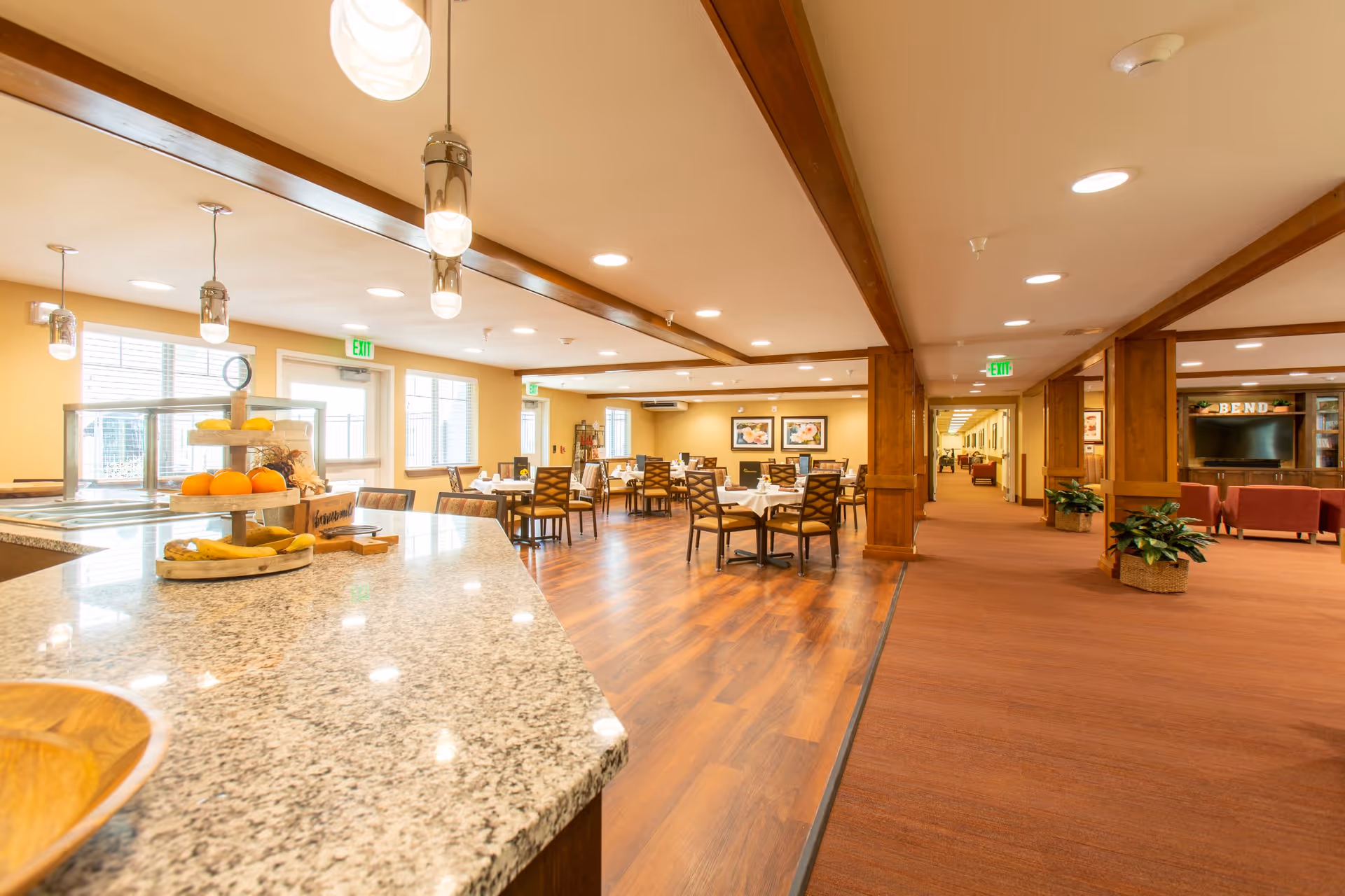 Bright and spacious dining area in a retirement community with wooden floors, tables and chairs set for meals, a granite countertop with fruit display, and warm lighting. The space extends into a hallway with carpet and seating areas, decorated with plants and framed artwork.