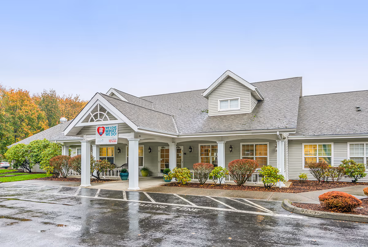 Exterior view of a single-story senior living facility building with a covered entrance supported by white columns. The building has gray siding, multiple windows with white trim, and a gabled roof. There are shrubs and small trees planted around the entrance, and the pavement in front is wet from rain.