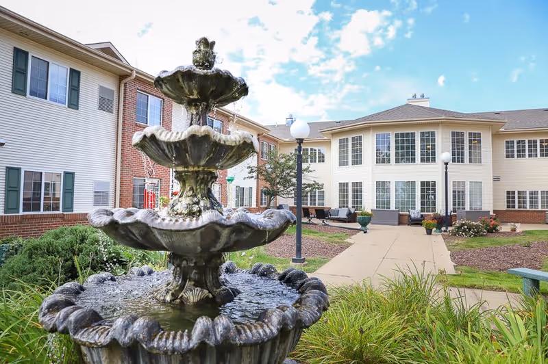 Outdoor courtyard area of a senior living facility featuring a three-tiered water fountain in the foreground, surrounded by greenery and plants. In the background, there is a two-story building with large windows, outdoor seating, and lamp posts under a partly cloudy sky.