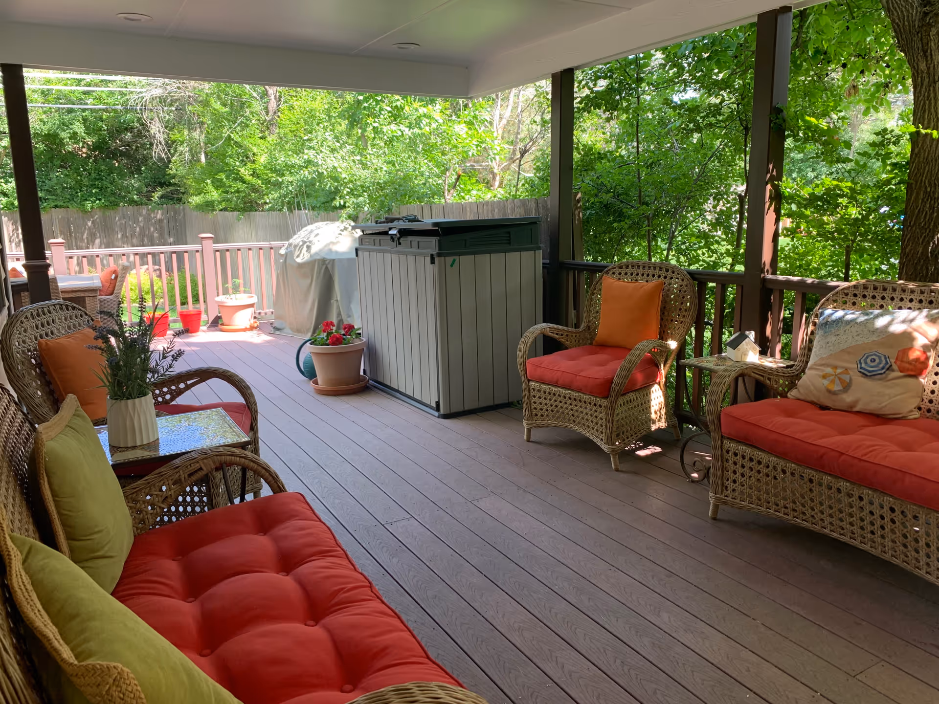 Covered outdoor patio with wicker furniture including a loveseat, two armchairs, and a side table. The furniture has red cushions and some pillows. There are potted plants and a covered grill on the wooden deck. Green trees and a wooden fence are visible in the background.