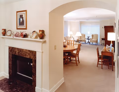 Open-plan interior showing a fireplace in the foreground and a dining table with chairs leading to a bright seating area with armchairs and lamps.