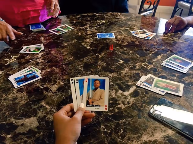 A group of people playing a card game around a dark marble-patterned table. One person is holding four cards, including one featuring Obi-Wan Kenobi. Other cards and a smartphone are visible on the table.