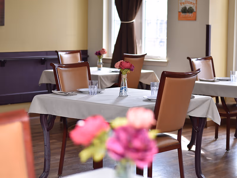 Small dining room with tables covered in white tablecloths, chairs, and vases of pink flowers.