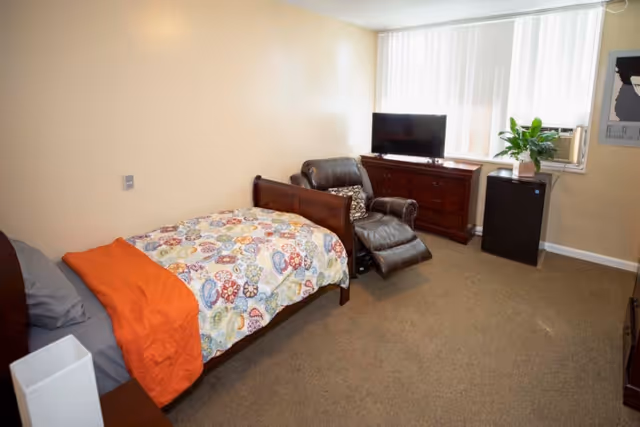 A senior living facility bedroom with a single bed covered in a colorful patterned blanket and an orange throw. Next to the bed is a brown leather recliner chair with a decorative pillow. Across from the chair is a wooden dresser with a flat-screen TV on top. A small black refrigerator and a potted plant are placed near a window with vertical blinds. The room has beige walls and carpeted floor.