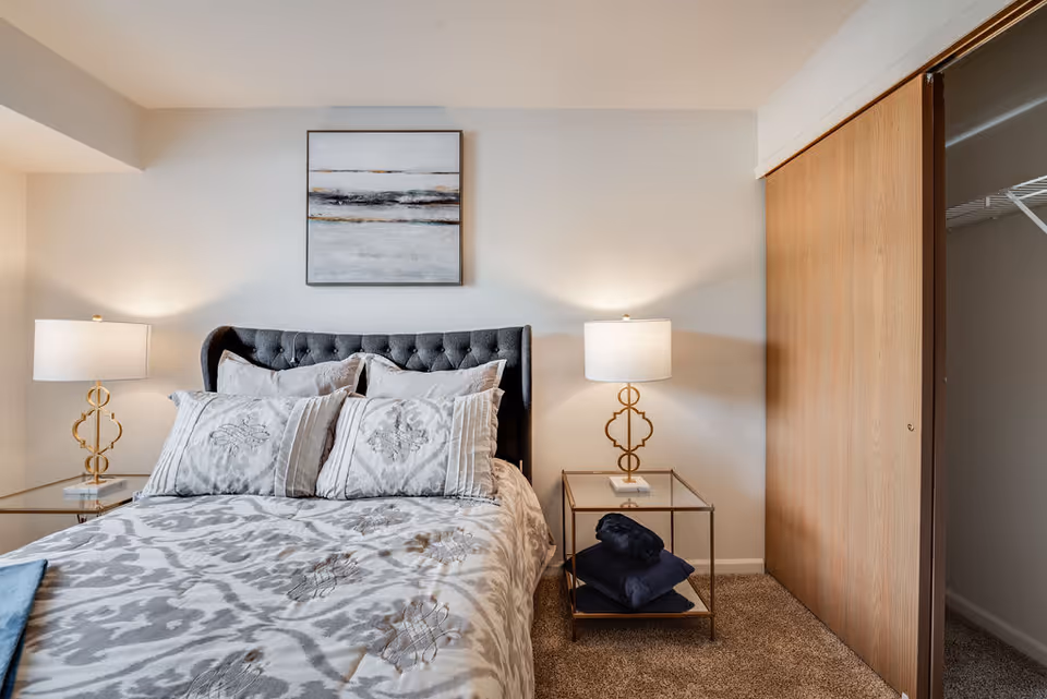 A neatly made bed with a tufted dark gray headboard, patterned gray and white bedding, and multiple pillows. On each side of the bed are glass-top nightstands with gold-colored lamps. A framed abstract painting hangs on the wall above the bed. To the right is a wooden sliding closet door, partially open to reveal a closet with a wire shelf. The room has beige carpet and neutral-colored walls.