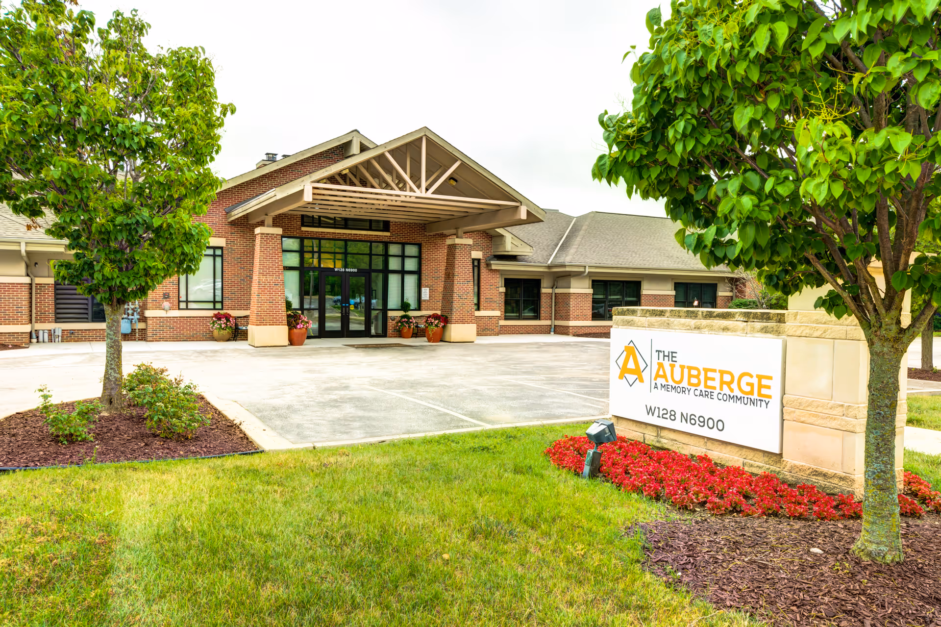 Exterior view of The Auberge at Oak Village, a memory care community building with a brick facade, large glass entrance doors, and a covered entryway. The foreground features a well-maintained lawn, two trees, flower beds, and a sign displaying the facility's name and address.