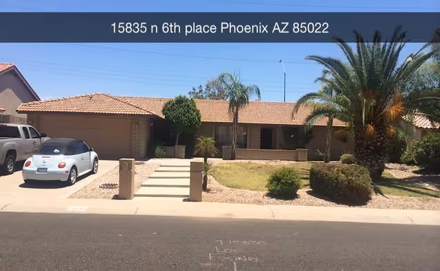 Single-story residential building with a tiled roof, a driveway with two parked cars, palm trees, and desert landscaping in front under a clear blue sky. The address 15835 n 6th place Phoenix AZ 85022 is displayed at the top of the image.