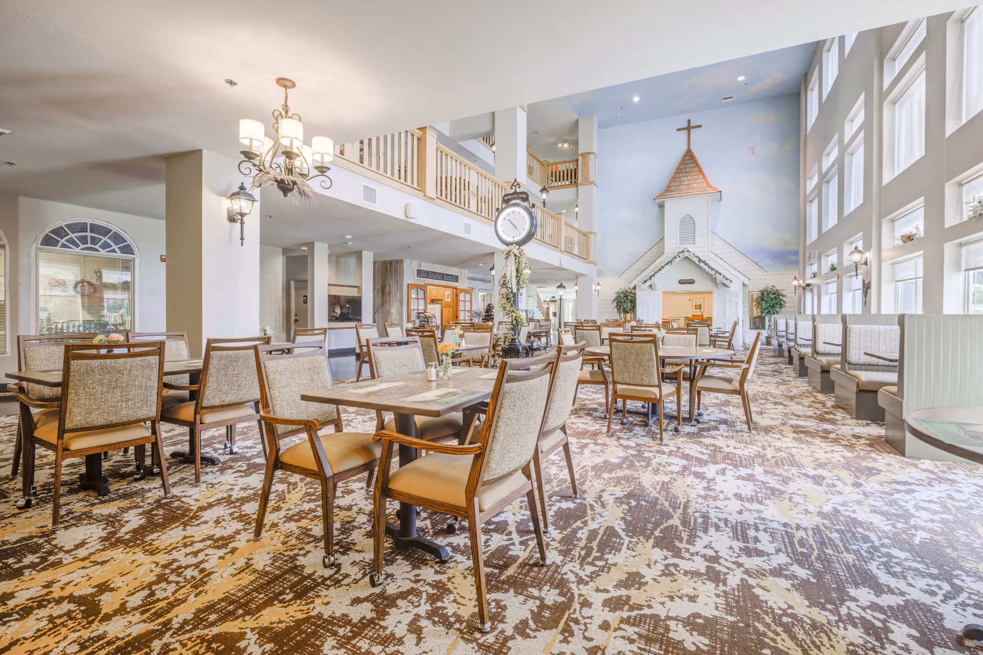 A spacious dining room with multiple tables and chairs arranged neatly. The room features large windows on the right side allowing natural light to fill the space. At the far end, there is a decorative structure resembling a small white chapel with a cross on top. The ceiling is high with a balcony railing visible above, and there is a large clock mounted on a post in the center of the room. The carpet has a patterned design in shades of brown and beige.