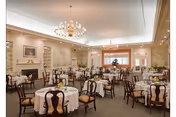 A spacious, elegant dining room with multiple round tables covered in white tablecloths, each set with glassware and napkins. The room features classic wooden chairs, chandeliers hanging from the ceiling, and soft lighting. There are built-in shelves and framed artwork on the walls, creating a warm and inviting atmosphere.