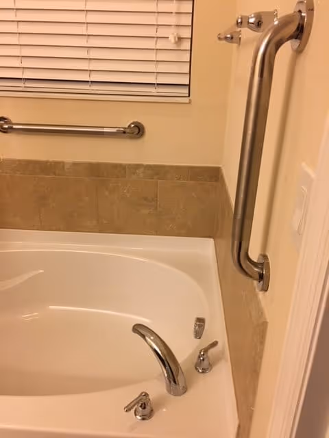 A bathtub with beige tiled walls and two stainless steel grab bars mounted on the walls, one horizontal and one vertical. The bathtub has a chrome faucet and handles. A window with closed white blinds is visible above the tub.