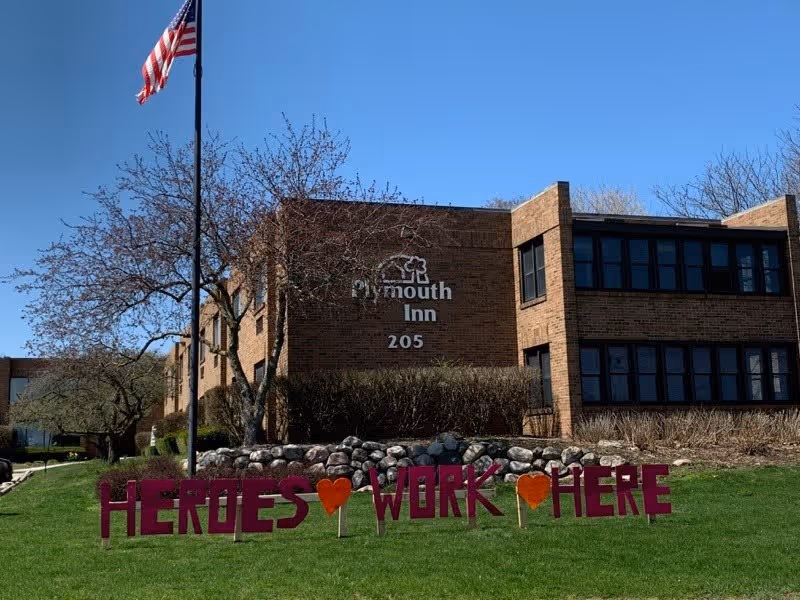 Exterior view of a brick building with the sign 'Plymouth Inn 205' on the wall. In front of the building, there is a green lawn with a sign made of large letters and hearts that reads 'HEROES WORK HERE'. An American flag on a flagpole is also visible, along with leafless trees and a clear blue sky.