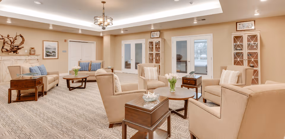 A spacious and well-lit living room area with beige walls and carpeted floor featuring a geometric pattern. The room is furnished with beige armchairs and sofas arranged around circular wooden coffee tables, each decorated with a vase of white tulips. There are wooden side tables next to the chairs, a decorative wooden sculpture on a white mantel, framed artwork on the walls, and glass-front cabinets flanking a set of double doors with windows.