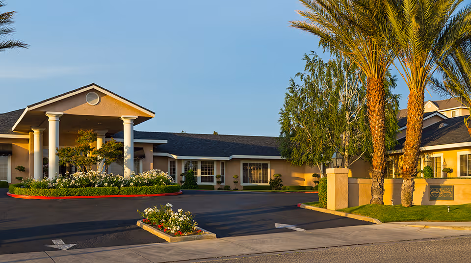 Exterior view of Sterling Commons Memory Care Community building with a driveway, landscaped garden beds with flowers, palm trees, and a clear blue sky.
