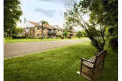 A peaceful outdoor scene at Spring House Estates featuring a stone building in the background, a paved road, green grass, trees, and a wooden bench in the foreground.