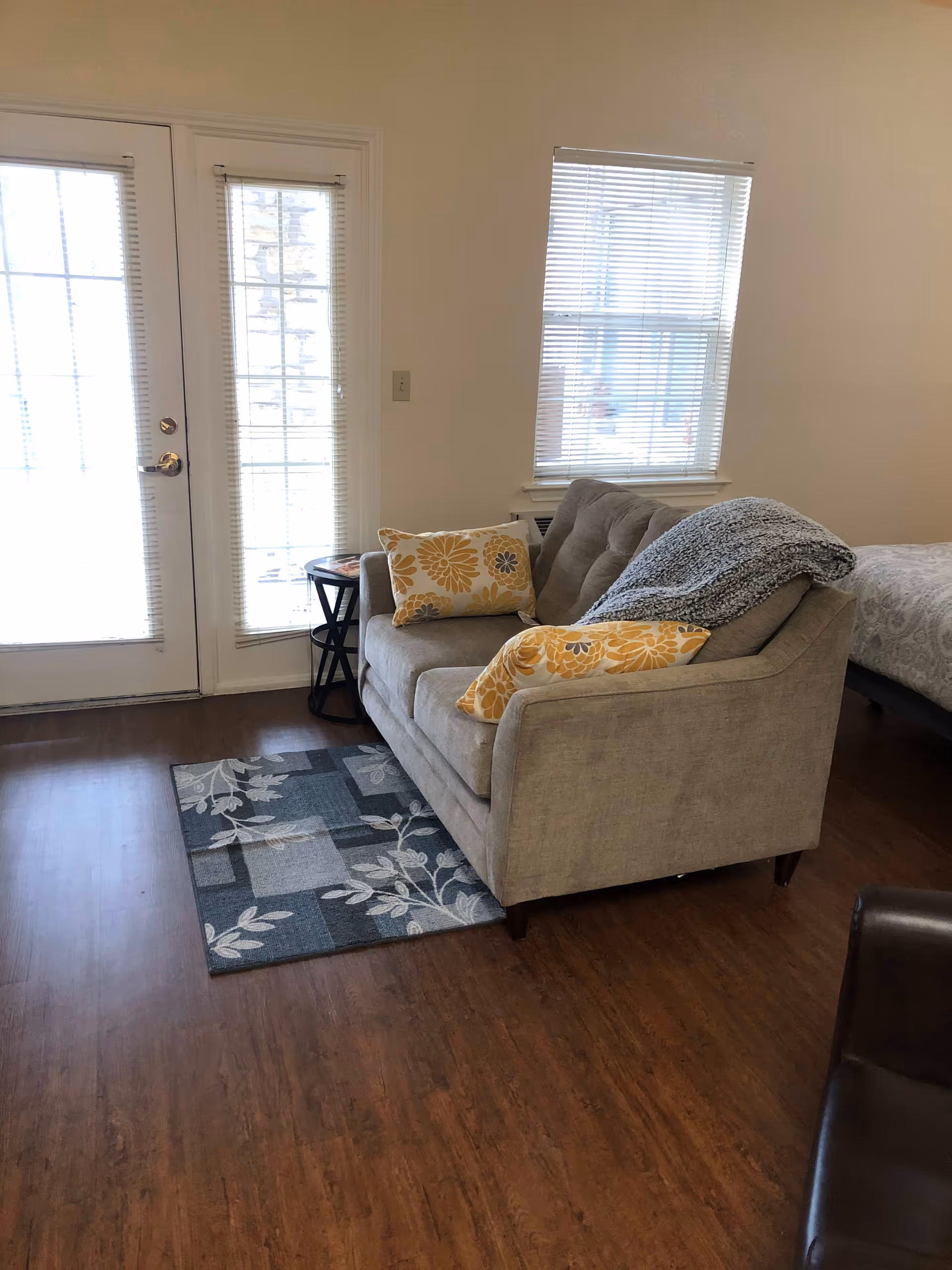 Cozy living area with a gray loveseat topped with yellow floral pillows and a throw, next to a window and glass door.