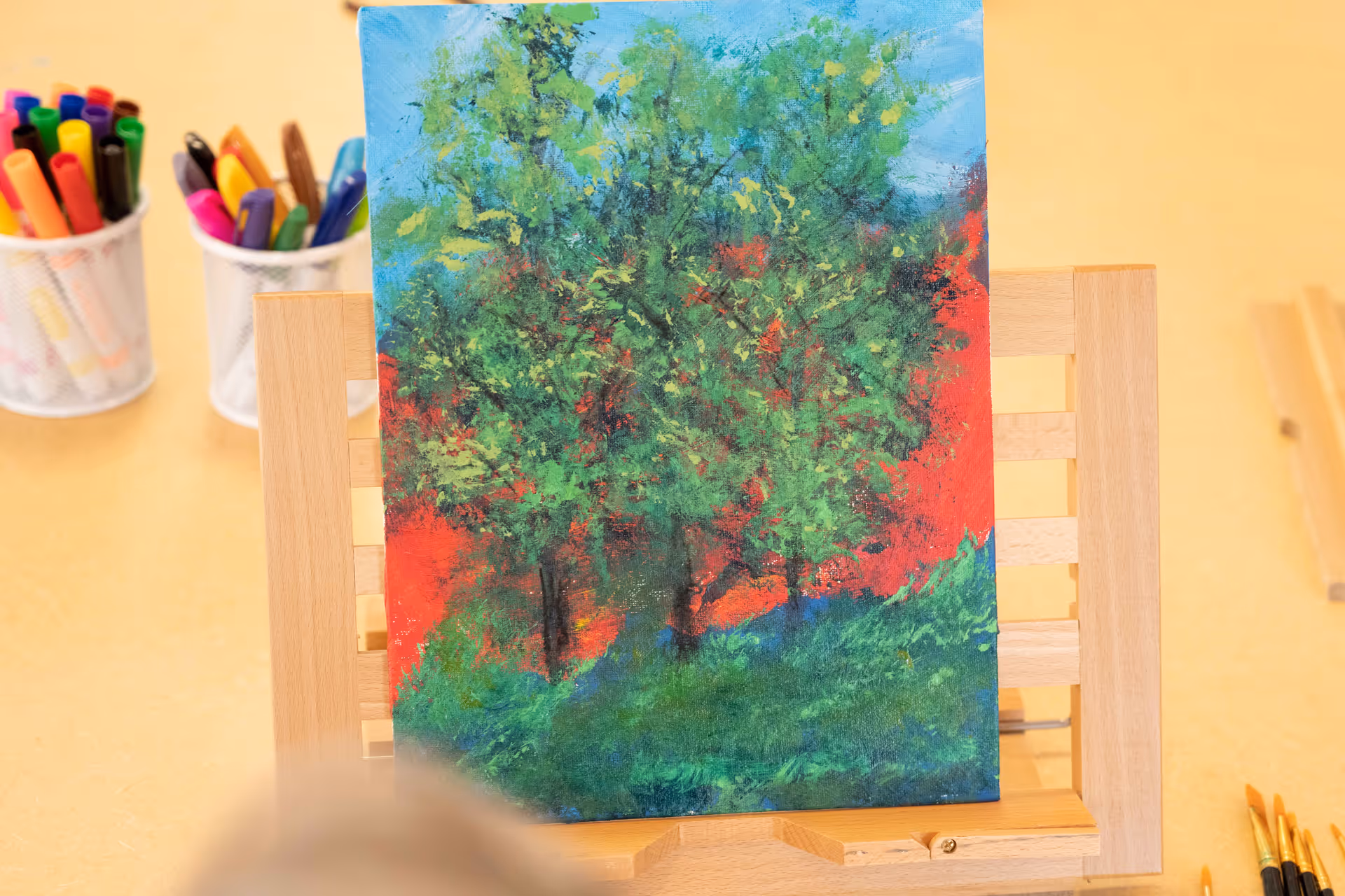 A colorful painting of green trees with a red background on a wooden easel, surrounded by art supplies including paintbrushes and markers in containers.