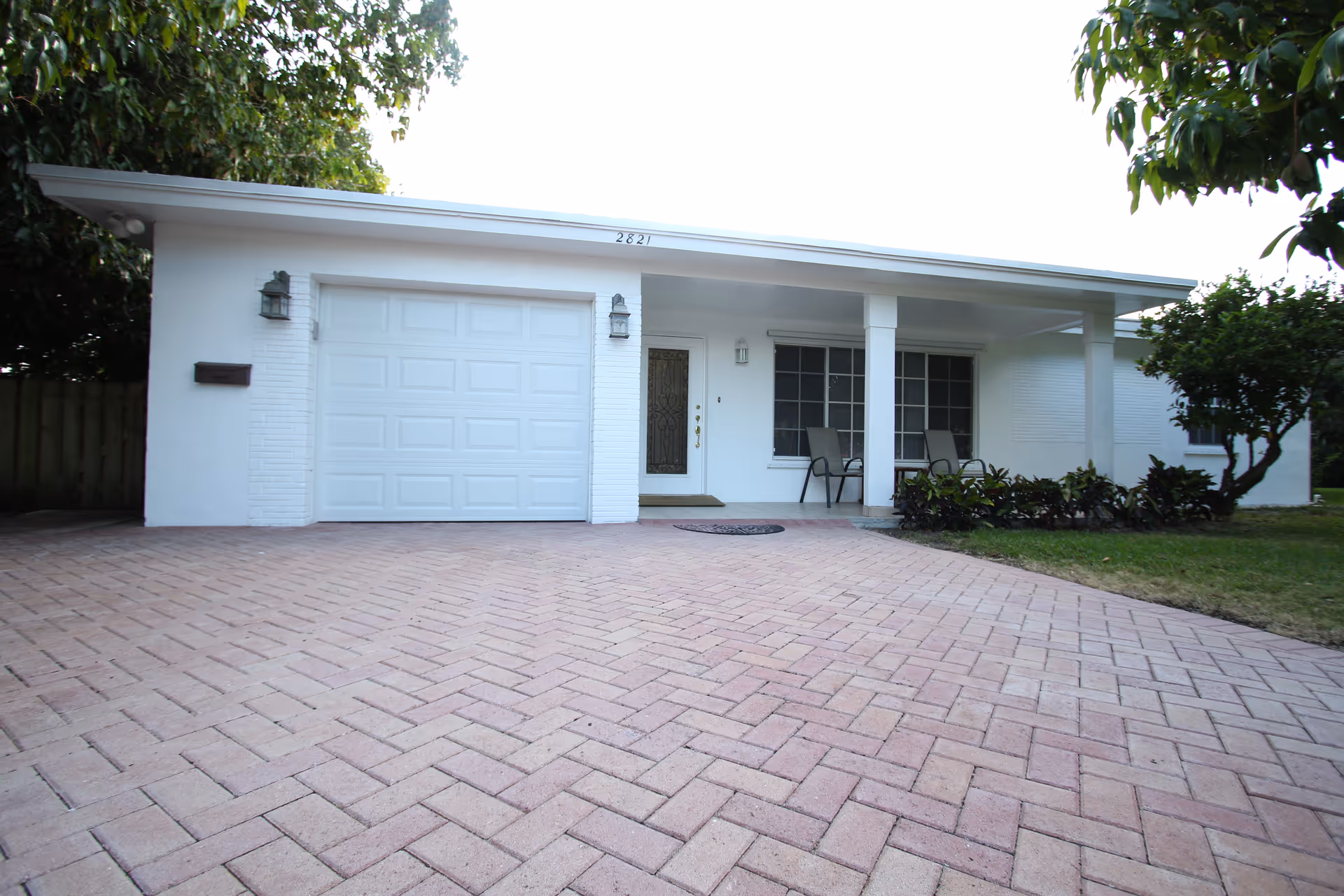 Front exterior of a single-story white house with an attached garage, covered porch with chairs, and a brick-patterned driveway.