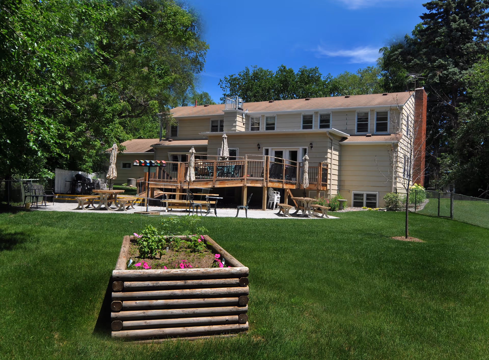 Backyard view of a two-story beige house with a wooden deck featuring tables, chairs, and closed umbrellas. The yard has a green lawn with a raised garden bed containing flowers and plants, surrounded by trees and a clear blue sky.