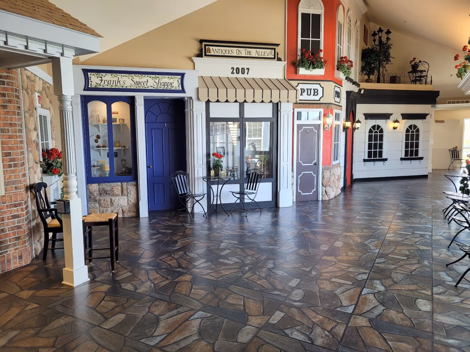 Indoor hallway designed to look like a small village street with storefronts including Frank's Sweet Shoppe, Antiques On The Alley, and a Pub. The floor is tiled with a stone pattern, and there are small tables and chairs placed along the walkway. The walls have decorative windows with flower boxes and lantern-style lights.