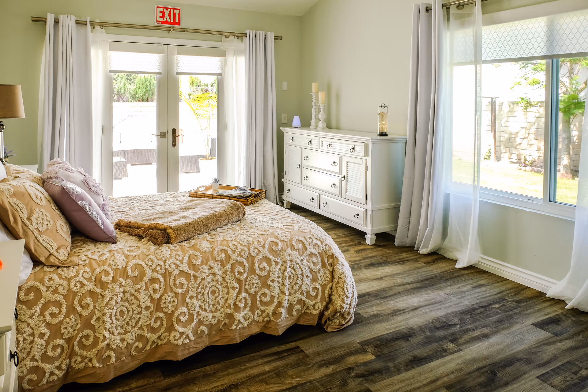 A bright bedroom with a large bed covered in a beige and white patterned comforter and multiple pillows. A folded brown blanket is placed on the bed along with a tray holding a small plant and a newspaper. To the right, there is a white dresser with decorative candles and a lantern on top. The room has large windows with sheer white curtains, and glass doors leading outside with an exit sign above them. The floor is wooden with a dark finish.