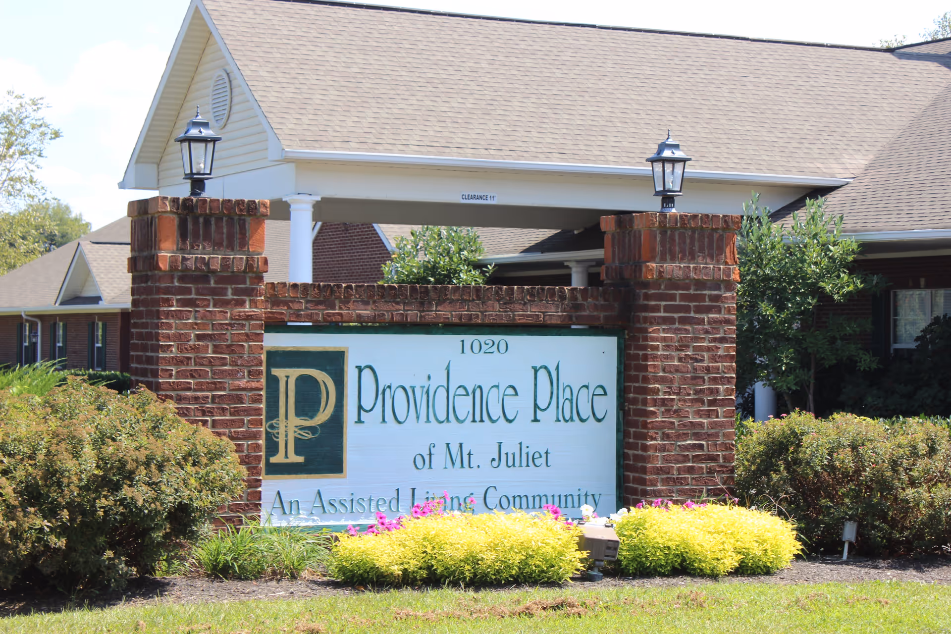 Entrance sign for Providence Place of Mt. Juliet, an assisted living community, set between two brick pillars with lanterns on top, surrounded by bushes and flowers with a building in the background.