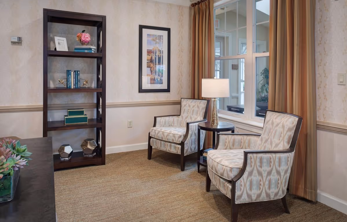 Two upholstered armchairs flank a small round side table with a lamp beside a window, with a bookshelf and framed art in a cozy seating area.