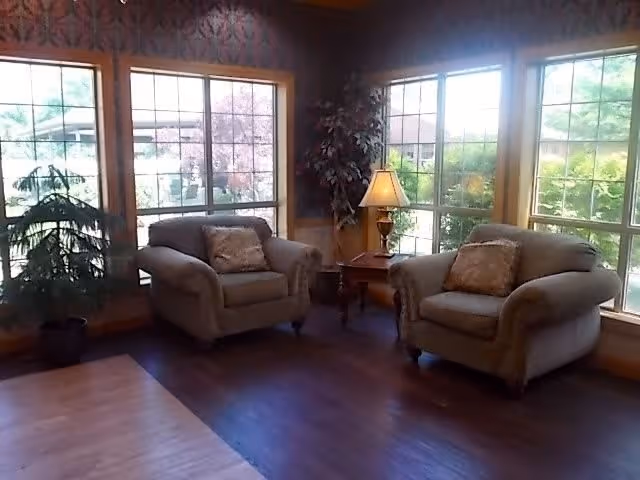 A cozy sitting area with two cushioned armchairs, each with a decorative pillow, positioned near large windows that let in natural light. There is a small wooden side table between the chairs with a lamp on it, and two potted plants are placed near the windows. The floor has a combination of wood and laminate flooring.