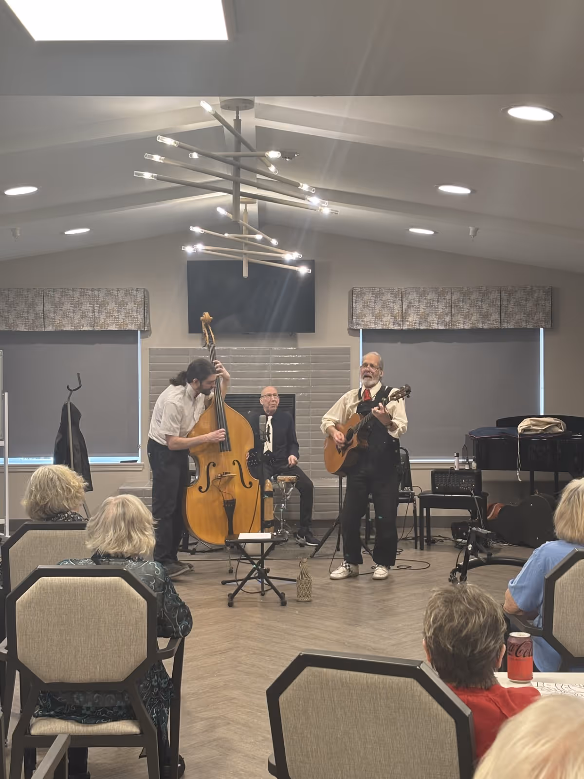 A small musical performance in a senior living facility common room. Three musicians are playing: one on a double bass, one on an acoustic guitar, and one seated behind them. Several elderly audience members are seated facing the performers. The room has modern lighting, a fireplace, and large windows with blinds.