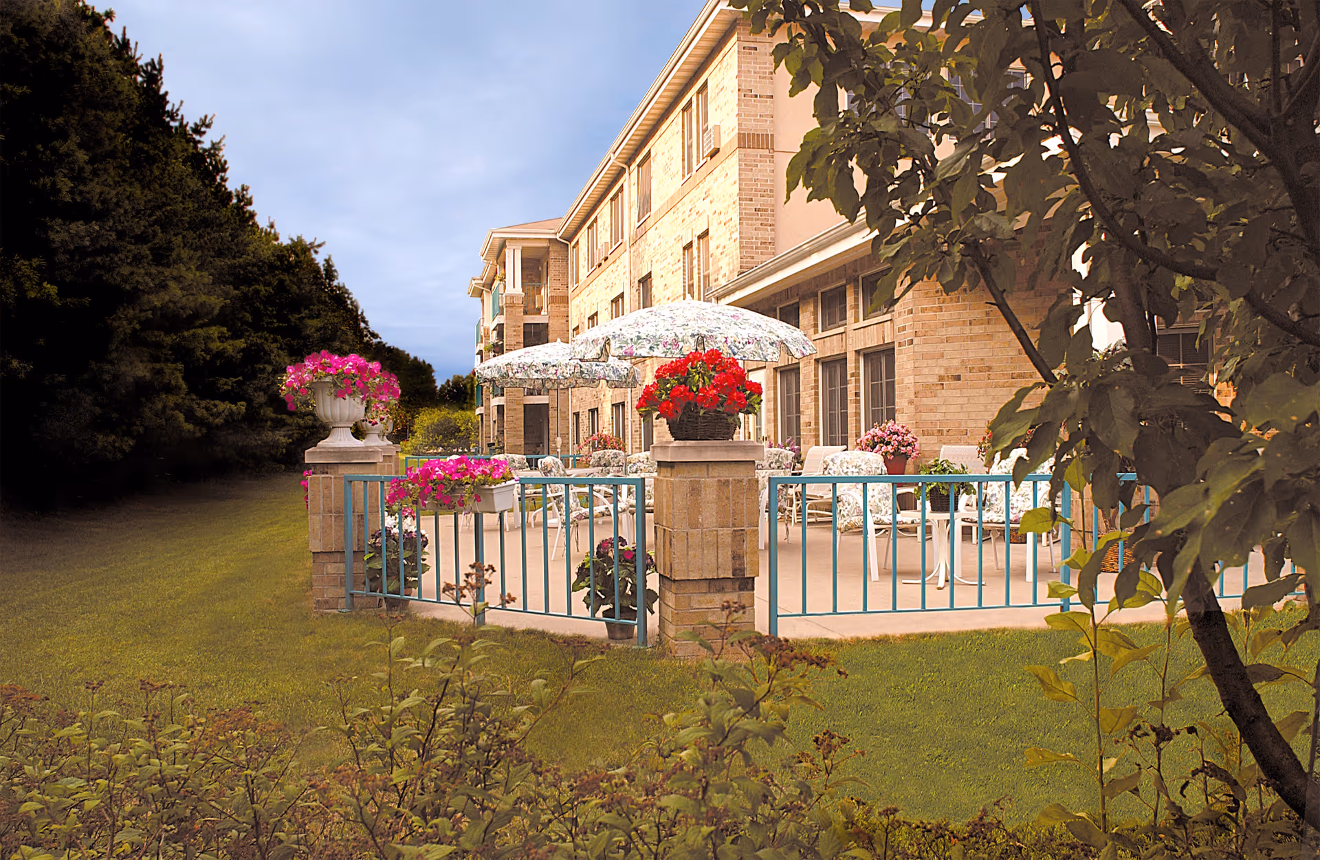 Outdoor patio area at Mulberry Glen with floral umbrellas, cushioned chairs, and potted flowers surrounded by a low blue railing. The patio is adjacent to a brick building with multiple windows, and there is green grass and trees around the patio.