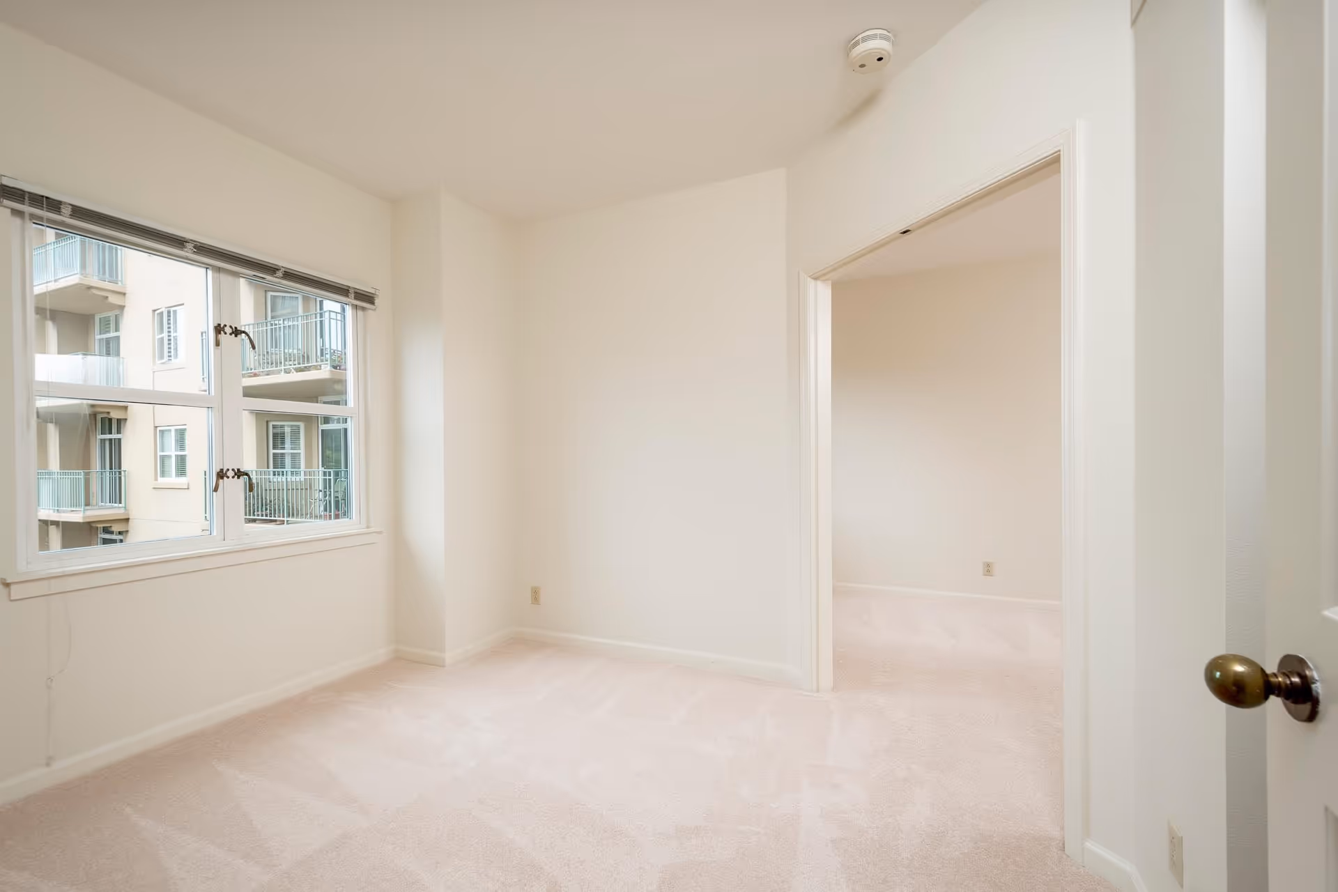 Empty bright bedroom with light carpeting, a large window showing neighboring balconies, and an open doorway to an adjacent room.