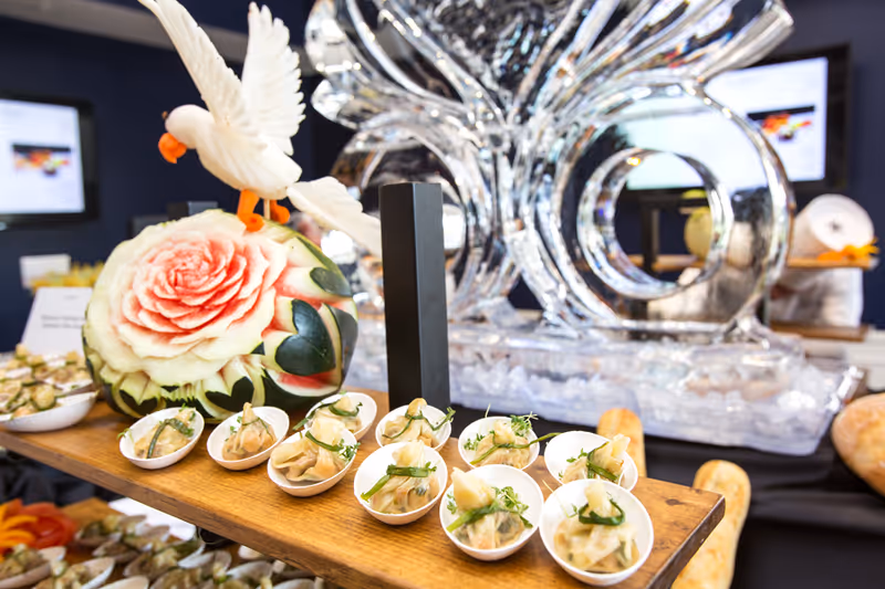 A close-up view of a wooden serving board with small appetizer dishes arranged on it, featuring dumplings garnished with herbs. Behind the board, there is an intricately carved watermelon with a rose design and a white bird sculpture on top. In the background, a large decorative ice sculpture is visible along with blurred screens and other food items.