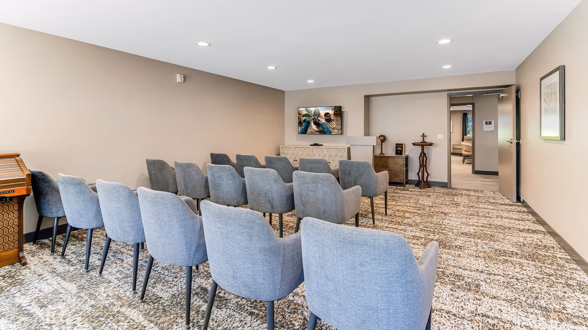 A small meeting or presentation room with three rows of gray upholstered chairs facing a wall-mounted flat screen TV. The room has beige walls, a patterned carpet, and recessed ceiling lights. There is a wooden cabinet and a small table with decorative items near the TV, and an open door leading to another room.