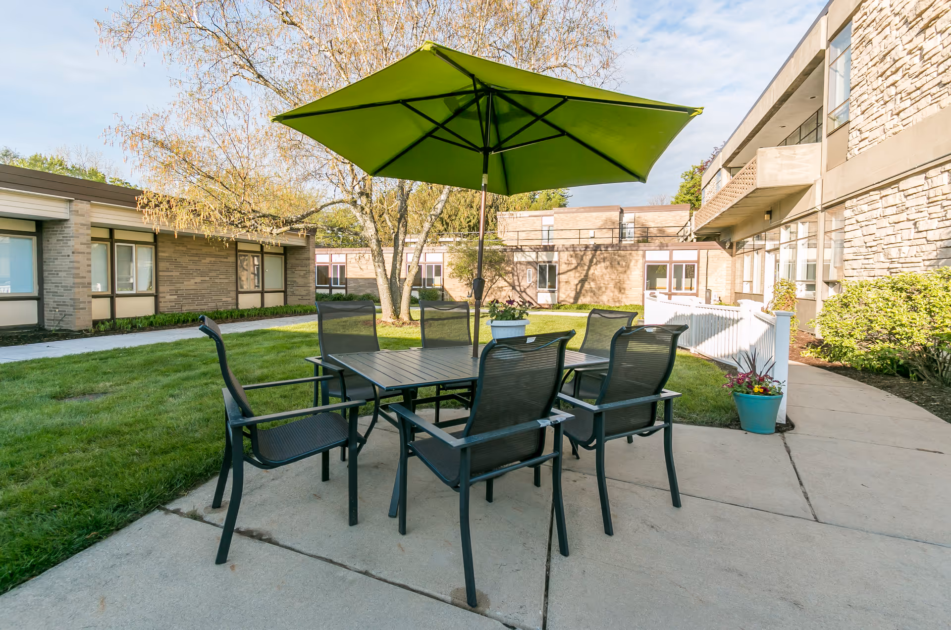 Outdoor patio area with a black metal table and six matching chairs under a large green umbrella. The patio is surrounded by grass and buildings with brick and stone exteriors. There are potted plants near the patio and trees in the background.